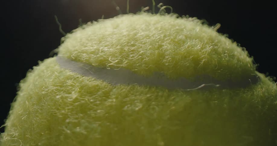 A macro closeup captures a bright yellow tennis ball as it slowly rotates against a solid black background