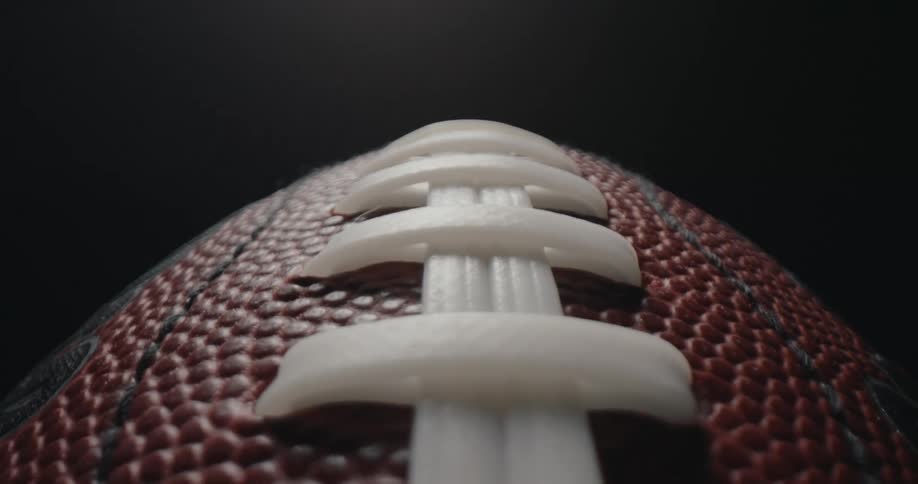 A macro camera shot slowly glides over the textured surface of an american football highlighting the white plastic laces and the pebbled brown leather against a darkt background