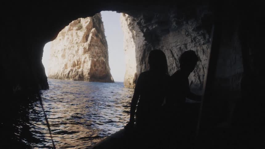 A man and a woman sit in a small boat as it floats through a dark sea cave and the couple is silhouetted against the bright sunlight at the cave entrance looking out at a large vertical rock formation rising from the blue water outside