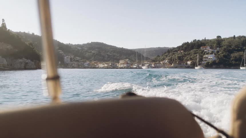 A view from a boat shows the white wake of churned water as it travels away from a coastal village tucked into a lush green hillside under a bright morning sun