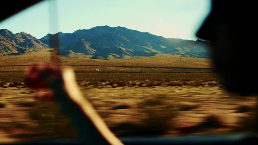 A young woman drives through the countryside as the wind blows her hair lifting one hand to adjust it while a mountain range stretches across the background