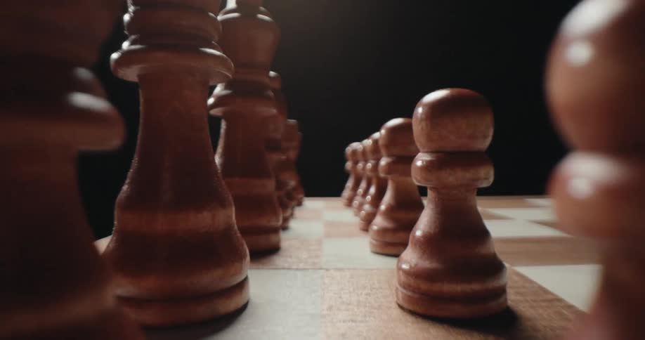 A close view of brown wooden chess pieces standing in two parallel lines on a checkered board under a bright light against a dark background