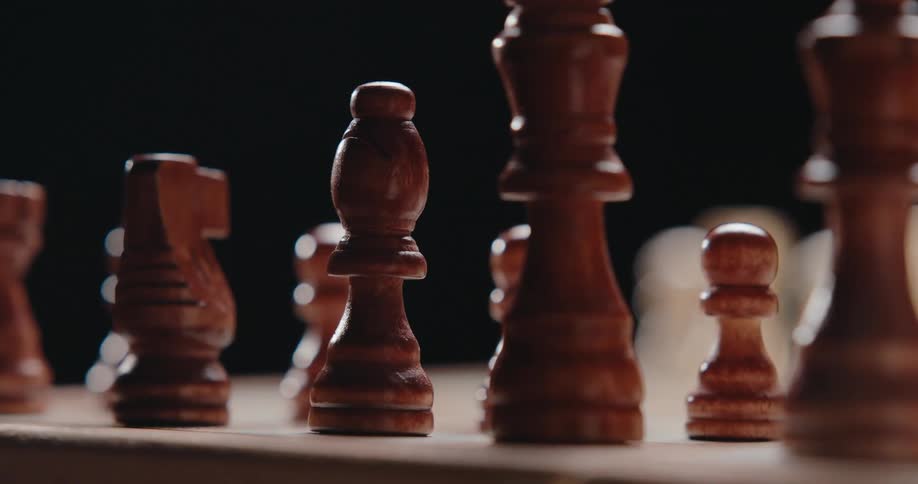 The camera pans across a wooden chessboard in a dark room capturing a row of brown chess pieces in shallow focus as they blur in and out of view