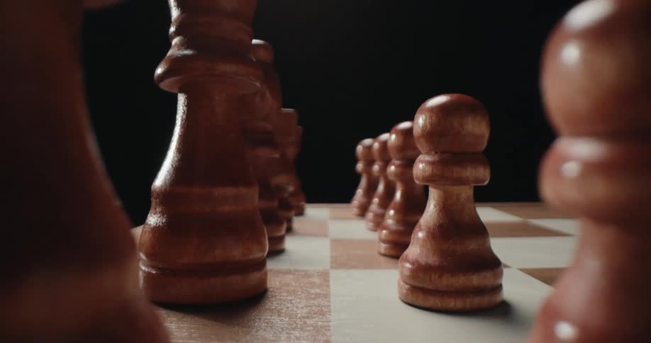 A lowangle tracking shot moves forward between rows of wooden chess pieces lined up on a board against a dark background