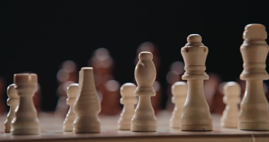 A camera pans across a wooden chessboard moving past white pieces including a bishop queen and king while brown pieces remain out of focus in the dark background