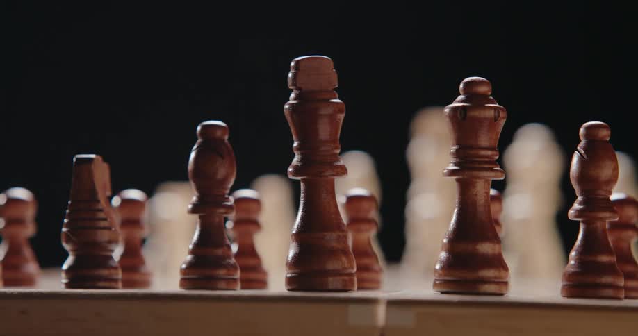 The camera pans across a wooden chessboard showing a row of dark brown chess pieces in sharp focus while lightcolored pieces remain blurred in the background against a dark setting