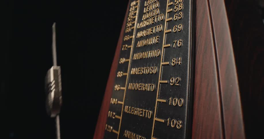 A closeup of a wooden metronome shows its black tempo scale with gold markings and italian musical terms dramatically lit against a dark background