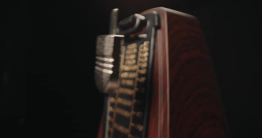 A mechanical wooden metronome with a silver pendulum swings steadily back and forth against a dark backgroundthe closeup shot focuses on the rhythmic movement of the weight as it passes the numbered tempo markings on the face of the device