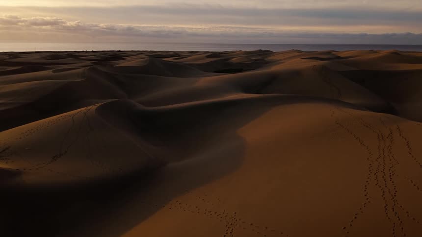 An aerial camera glides over vast sand dunes marked with footprints under a sunset sky showing the desert meeting the distant ocean horizon as long shadows stretch across the textured landscape