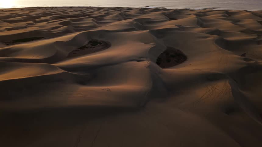 The video features an expansive aerial view of undulating sand dunes that stretch across the landscape during a golden hour sunset in the distance the desert terrain meets the calm waters of the ocean under a soft hazy sky