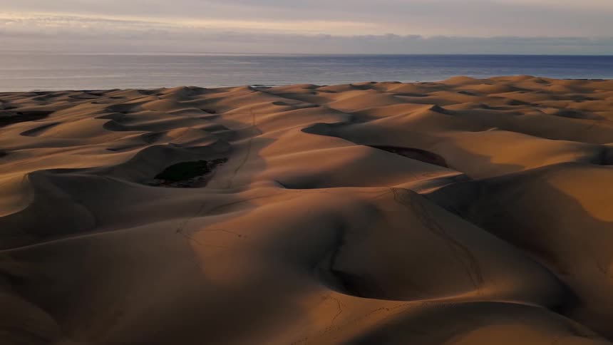 An aerial drone shot glides over expansive sand dunes toward the ocean under a hazy sky