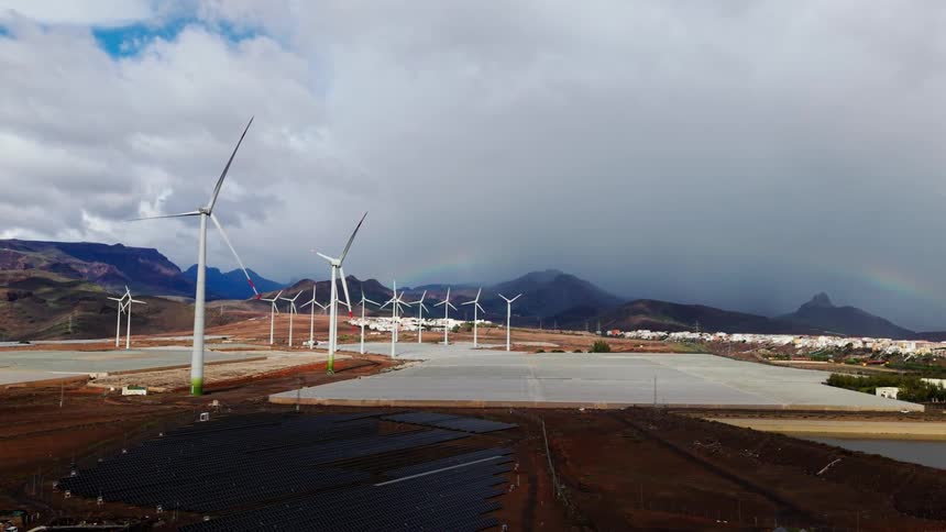 Rows of tall white wind turbines turning across a reddish open landscape backed by distant mountains and dramatic storm clouds