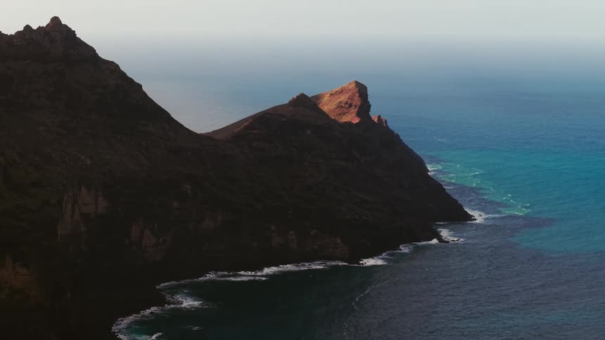 Aerial footage shows a sharp volcanic mountain ridge descending into a turquoise ocean where white waves crash against dark rocky cliffs under clear daylight