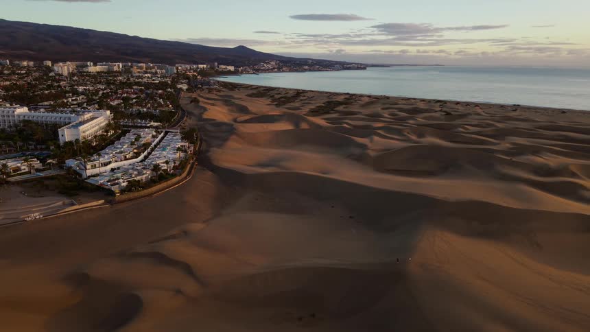 An aerial view shows a vast expanse of rolling sand dunes meeting a coastal town with white buildings and a calm blue ocean under a clear sky during sunset