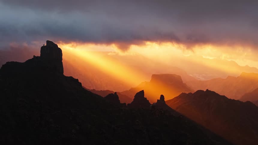 Sunlight pierces through a thick layer of clouds over a rugged mountain range at dusk with the sun rays illuminating the hazy valleys while the foreground rock formations remain in deep silhouette
