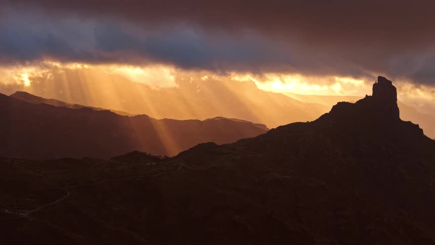 Sun rays pierce dense clouds and illuminate silhouetted mountains