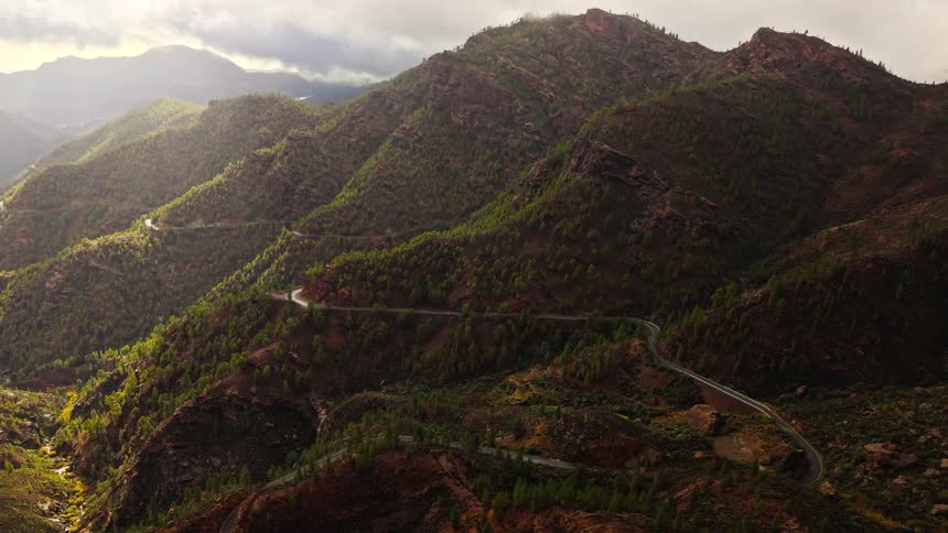 Aerial view of a winding road through steep tree covered mountains under a cloudy sky
