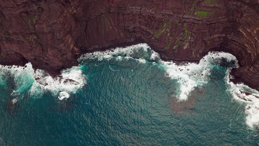 An aerial topdown view shows blue ocean waves crashing against the base of dark steep rocky cliffs topped with patches of green moss as white sea foam swirls around the jagged coastline