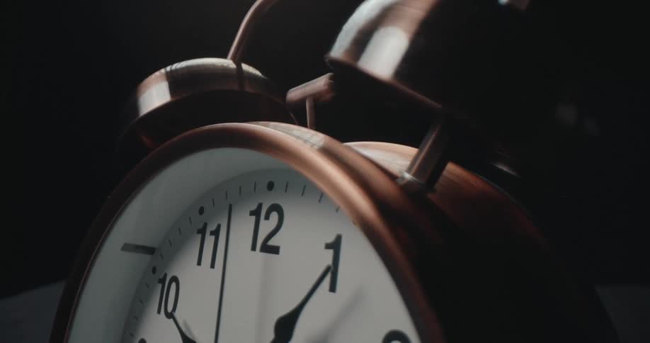 A copper twinbell alarm clock sits on a dark surface and rings loudly as the hammer rapidly strikes the bells causing the entire device to vibrate against a black background