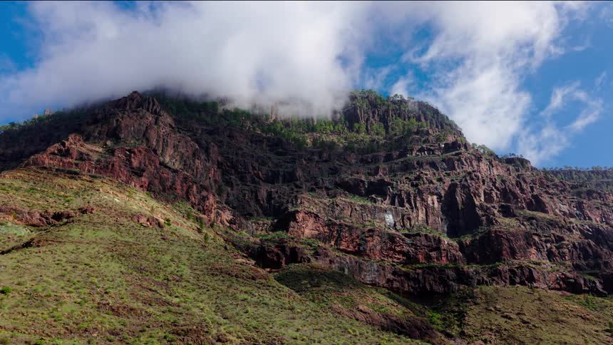 A massive rugged mountain cliff rises beneath a blue sky with drifting white clouds