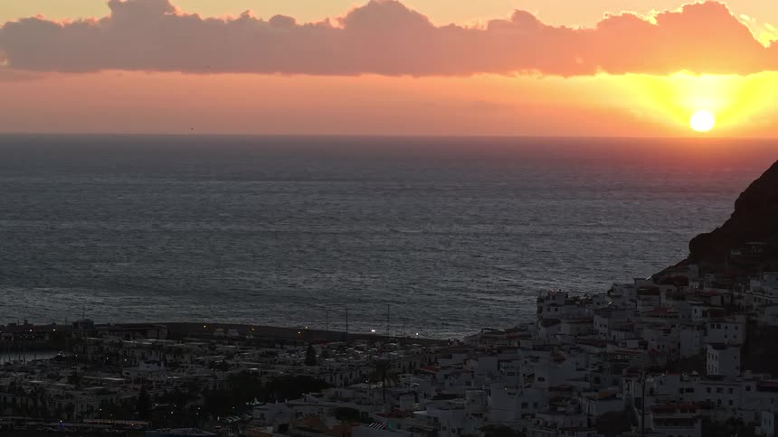 Golden sun sets over a calm ocean with a coastal town resting against a dark hillside