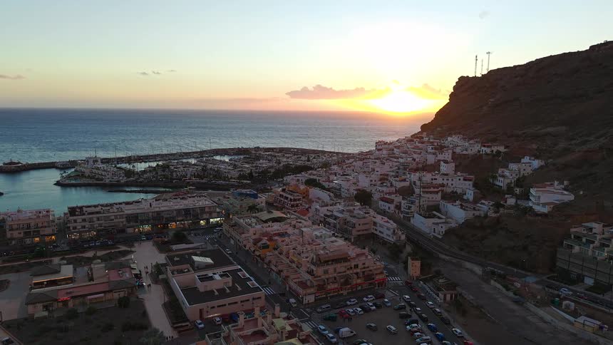 An aerial drone shot captures a coastal town at dusk showing white buildings clustered on a hillside next to a harbour while the sun sets over the ocean horizon