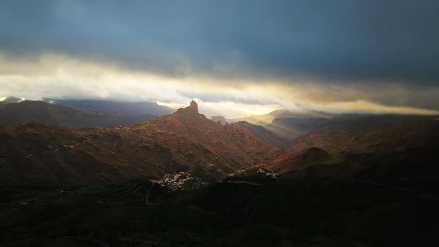 An aerial drone shot captures a wide mountain valley under a heavy cloud layer where sunlight breaks through to illuminate a prominent rock peak and a small cluster of white houses nestled on the green hillside