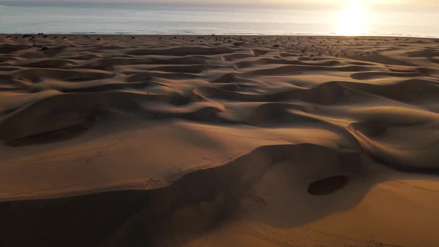 The camera pans over vast sand dunes at sunset toward the ocean as the low sun creates long shadows across the desert landscape
