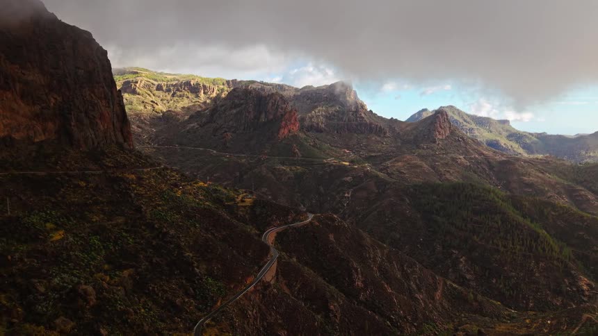 A winding mountain road curves through a vast canyon beneath a dramatic cloudy sky