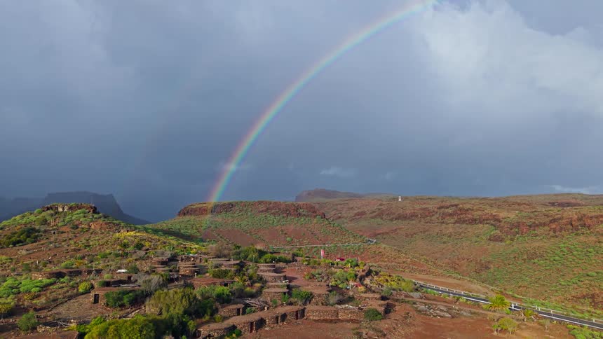 Vibrant rainbow spans cloudy sky above reddish hills and circular dwellings