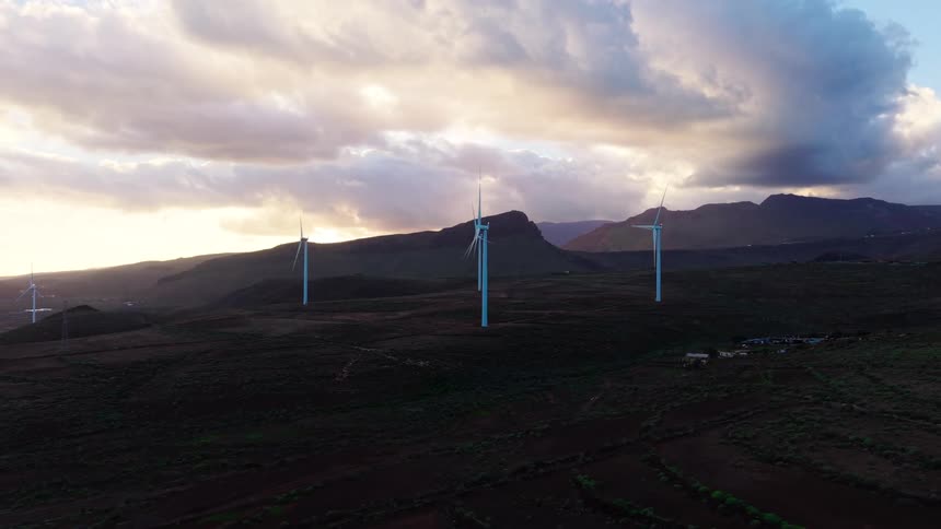 Wind turbines spread across elevated terrain with mountains in the background under a partly cloudy sky