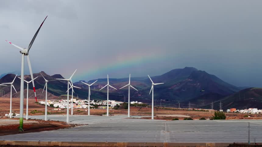 A row of wind turbines rises above a village and mountains under a stormy sky