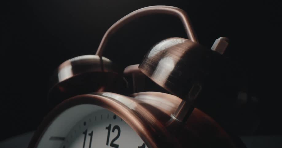 A close view of a copper twin bell alarm clock on a dark surface as its metal hammer strikes the bells rapidly against a black background