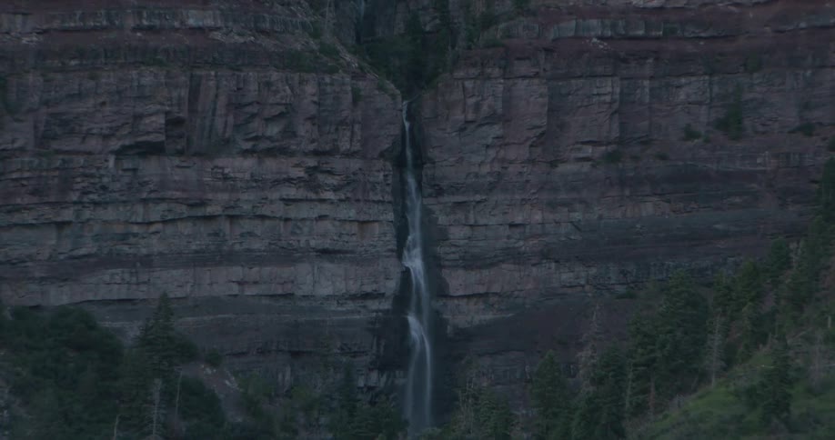 Narrow waterfall flows down a cliff towering over conifer covered slopes after dusk