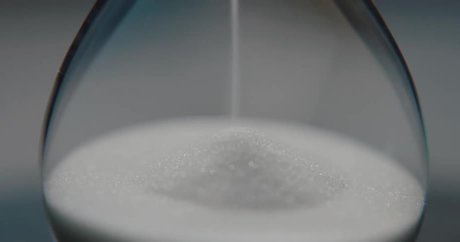 In an extreme closeup a steady stream of white sand falls and collects in the bottom of a glass hourglass marking the passage of time