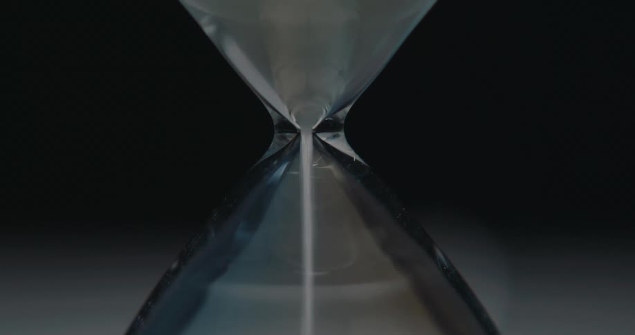 A close view shows white sand flowing steadily through the narrow center of a glass hourglass against a dark background