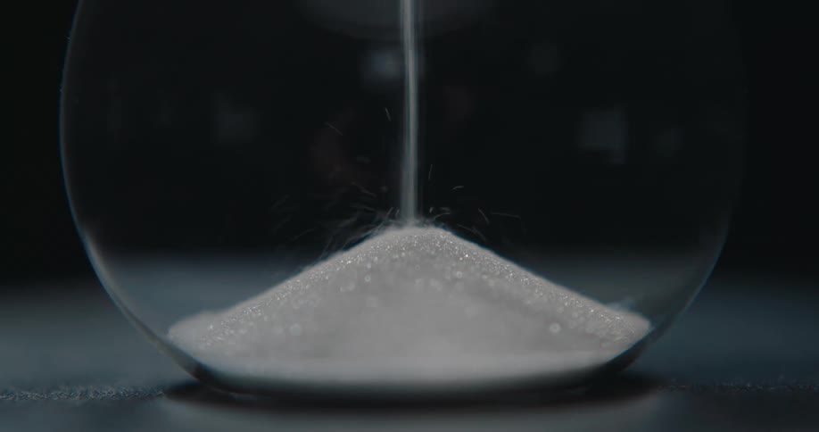 White sand falls steadily through a narrow neck into the bottom bulb of a glass hourglass forming a growing conical mound against a dark background