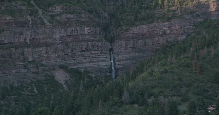 A thin waterfall cascades down a massive layered rock cliff surrounded by dense evergreen forests and steep mountain slopes