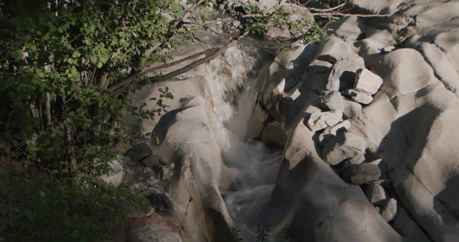 A small waterfall cascades down smooth lightcolored rock formations nestled within a shaded forest areathe camera zooms in closer to capture the rushing white water as it flows through the narrow worn channels of the stone