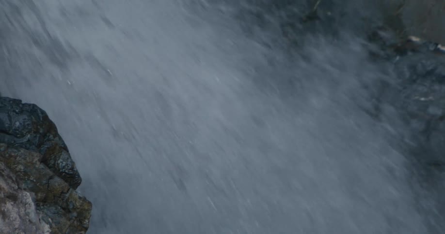 A camera zooms in toward a waterfall cascading down a narrow rocky gorge eventually filling the entire frame with the rushing white water as it pours between steep canyon walls and sparse vegetation