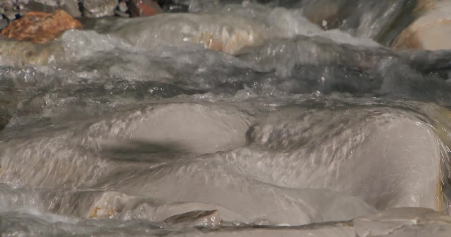 Clear water flows over smooth lightcolored rocks in a shallow mountain stream the camera zooms out to reveal a small waterfall surrounded by a tree trunk and forest debris under natural daylight