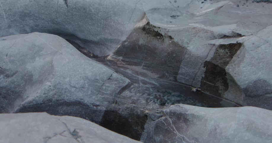 Clear water flows steadily through a narrow carved channel between smooth grey glacial rocks and ice formations creating small ripples as it moves downward across the uneven weathered terrain of the frozen landscape