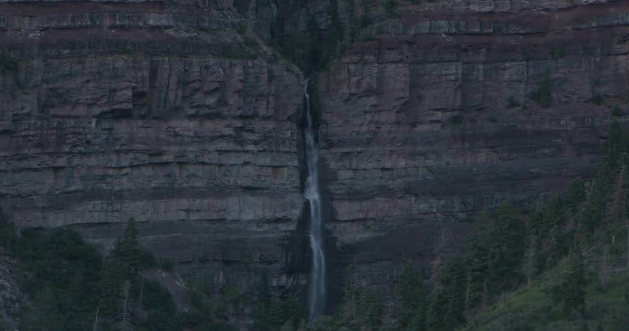 A slender waterfall threads down the center of a towering layered rock cliff its pale stream contrasting with the dark stone and dense evergreen forest below