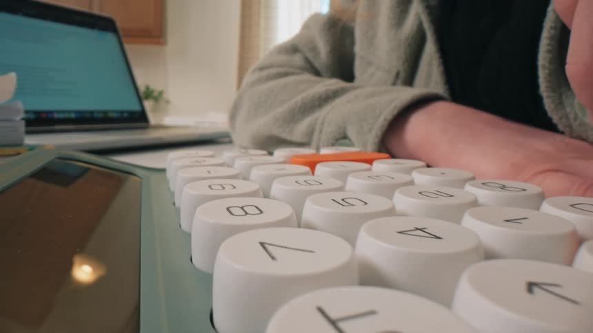 A woman wearing a grey fleece jacket uses a mechanical calculator with large round keys on a white desk next to a laptop and a blue pen while working in a bright room