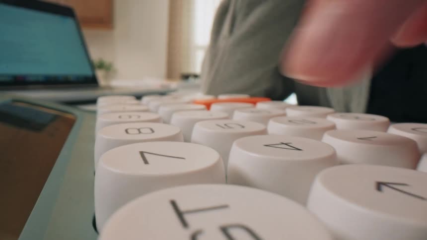 A persons hand presses large rounded buttons on a white and orange calculator in a closeup shot the camera then pulls back to show the person sitting at a desk with a laptop in the background