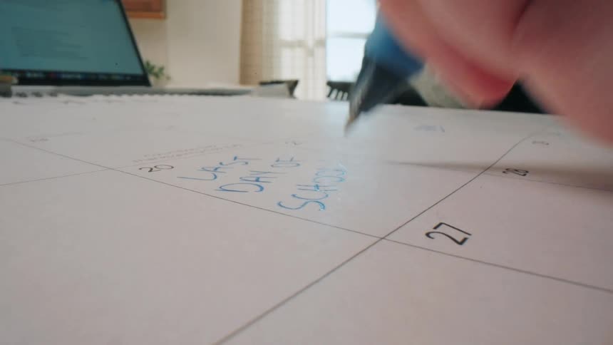 A person uses a blue pen to write the word school onto a calendar grid the camera is positioned at a low angle showing the tip of the pen moving across the paper while a laptop sits open in the background