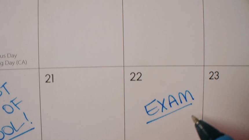 A closeup shot moves down a wall calendar showing handwritten notes like stephen foot surgery 9am and final the camera stops as a hand uses a blue pen to underline the word exam written on the 22nd