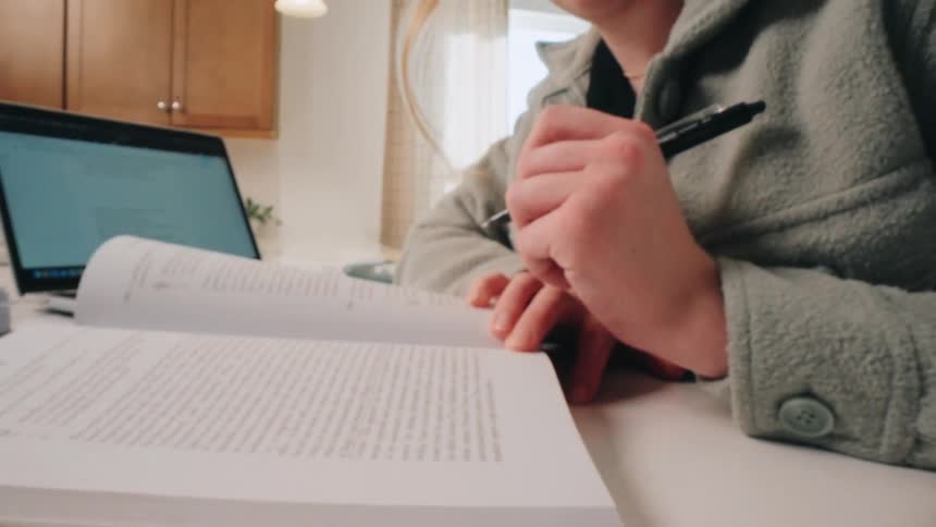 Person seated at a table writes in an open book with a laptop and study materials nearby