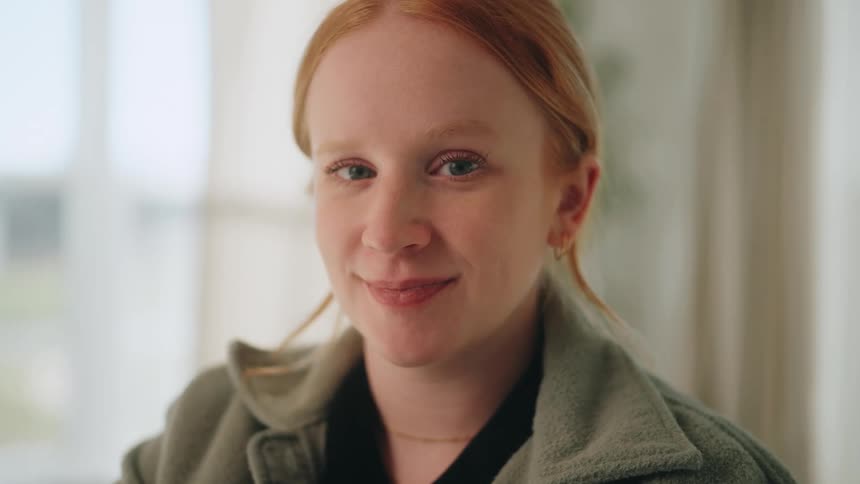 A young woman with red hair initially out of focus turns to the camera and comes into focus with a gentle confident smile