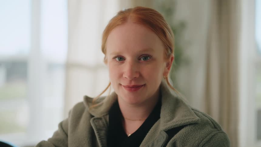 Redhaired woman in an indoor portrait with a neutral expression against a blurred background